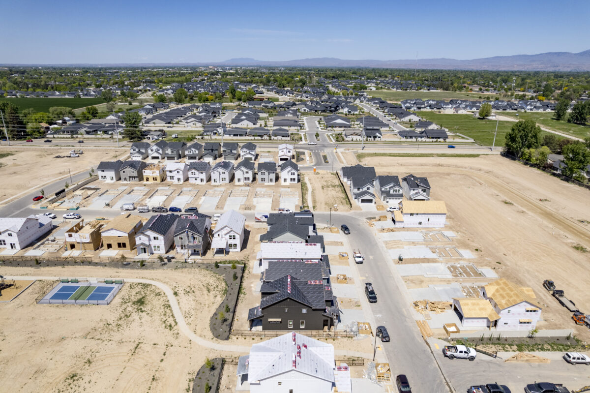 Drone view from the edge of the construction site looking toward an established neighborhood, with multiple new homes in framing and foundation stages in the foreground.