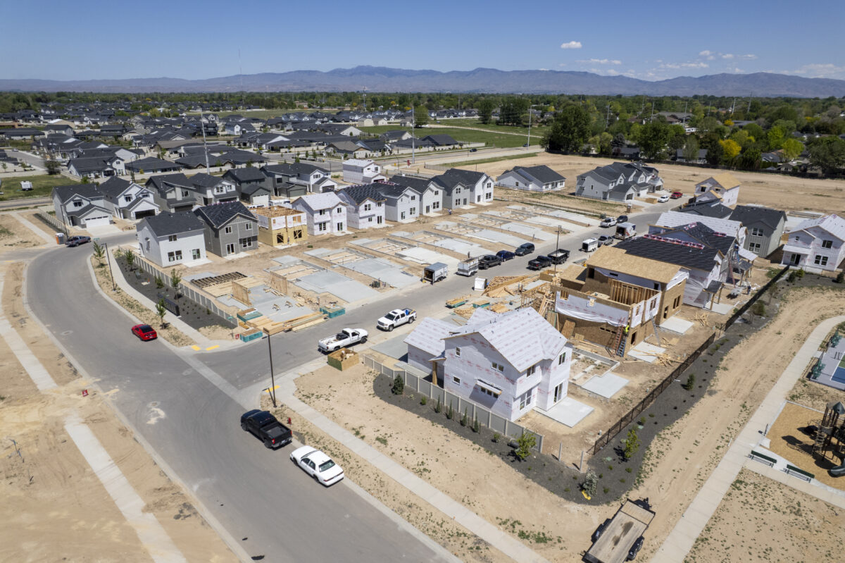 Overhead drone photo of a community build-out with rows of concrete foundations, multiple homes in framing stages, and finished houses lining the background under a blue sky.