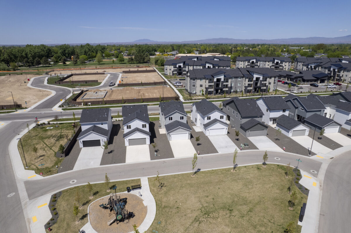 Elevated drone view showing a row of brand-new CBH Homes with clean modern designs and dark roofs, facing a small park with a playground. Behind the homes is a developing infill site with paved roads and utility groundwork in place, and a backdrop of mid-rise apartment complexes and distant tree lines under a clear blue sky.