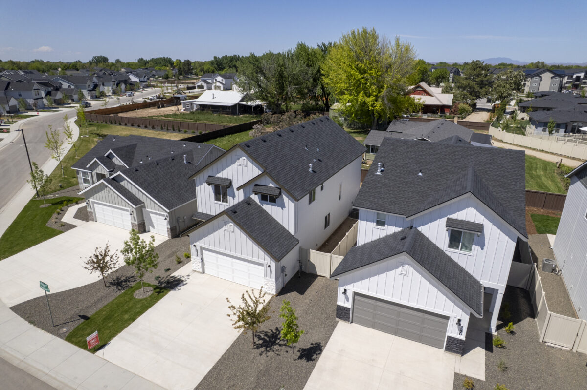 Close drone shot of modern two-story and single-story homes with white exteriors and dark roofs, sitting on tidy gravel and lawn landscapes along a freshly paved street.