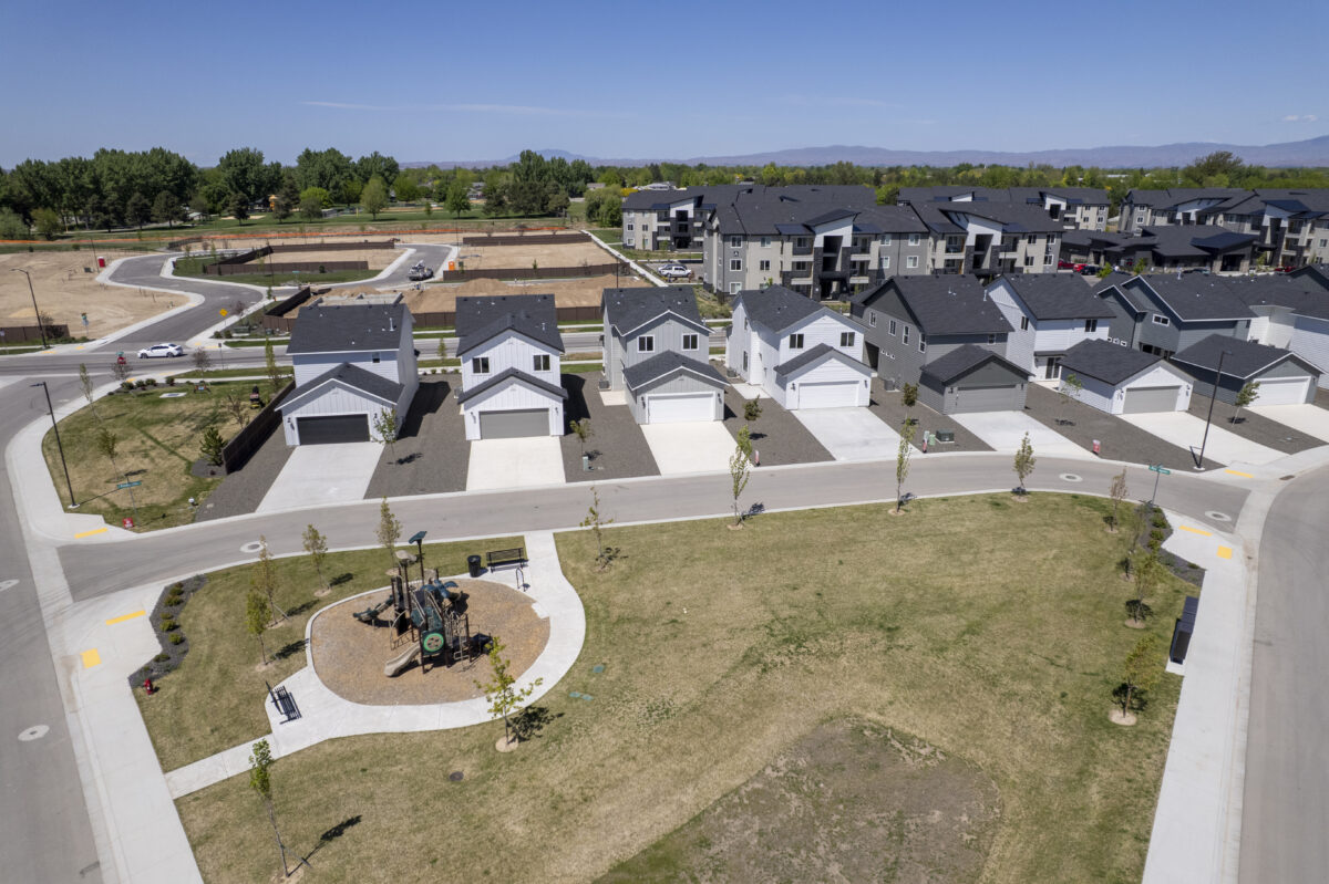 View from above a small community park with playground equipment, showing a row of uniform new homes directly across the street, with apartment buildings in the background.