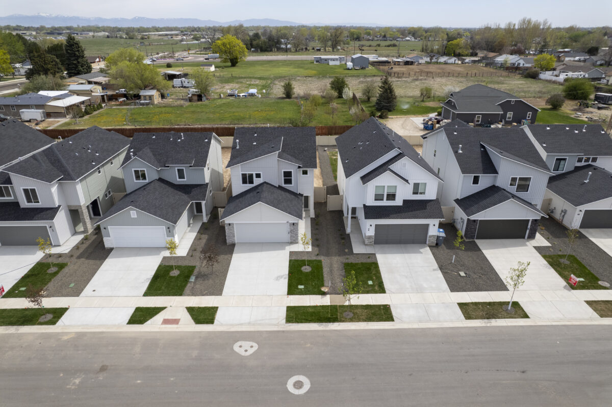 Aerial view of a row of newly constructed two-story suburban homes with dark roofs and attached garages, neatly aligned along a clean street with young trees and fresh landscaping. Behind the homes, a wooden privacy fence separates the development from open fields and rural properties, with mountains faintly visible in the distance under a cloudy sky.