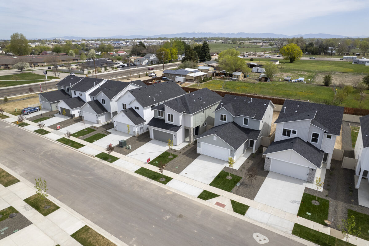 Angled aerial view of a residential street in the Walton Woods community, showcasing a variety of modern two-story homes with light-colored exteriors, black roofs, and small front lawns. The street runs parallel to a main road and rural farmland beyond, with green pastures and low buildings stretching out toward the base of distant mountains.