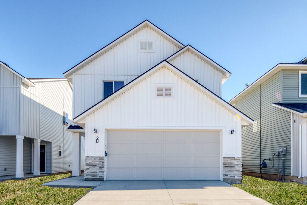 Front-facing view of a two-story home with clean white vertical siding, a double gabled roofline, and a two-car garage featuring a light gray door. Stone accents line the base of the exterior, and the home is framed by neighboring houses. The driveway and front lawn are well-maintained under a clear blue sky.