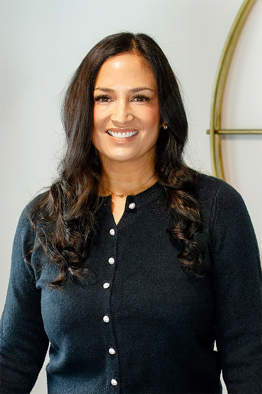 Professional headshot of a smiling woman with long dark hair, wearing a black button-up sweater, standing in front of a light background.