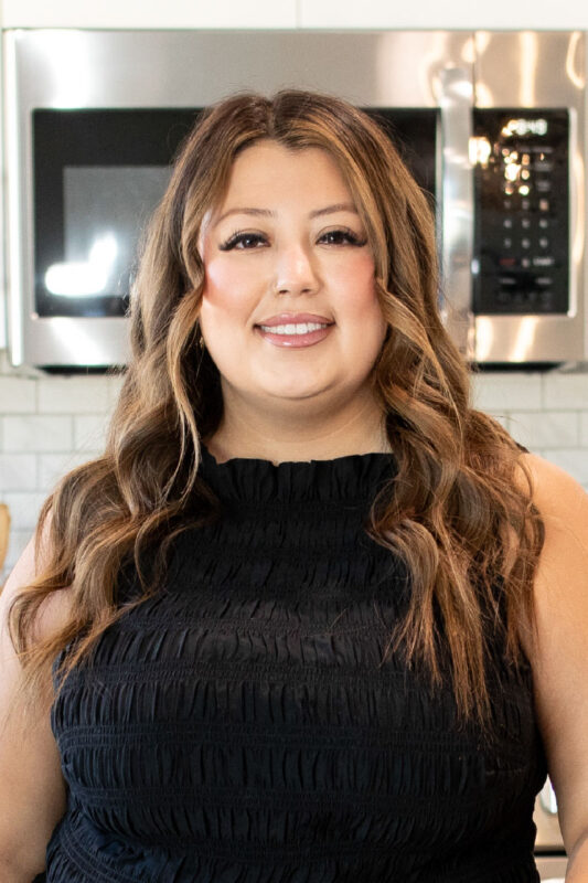 Professional headshot of a smiling woman with long wavy brown hair, wearing a sleeveless black textured top, standing in a modern kitchen with stainless steel appliances and white tile backsplash.