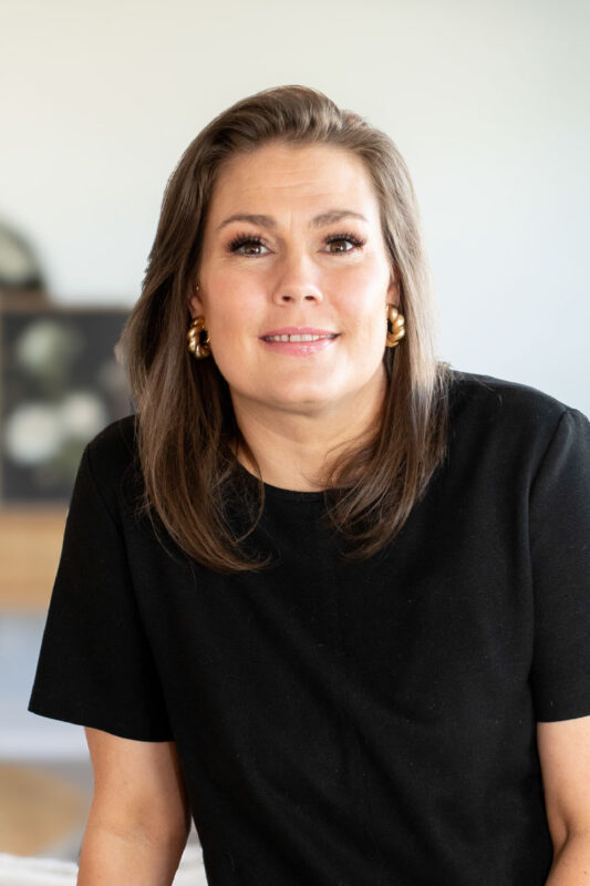 Professional headshot of a confident woman with straight shoulder-length brown hair, wearing a black top and gold hoop earrings, posing indoors with a soft, modern background.