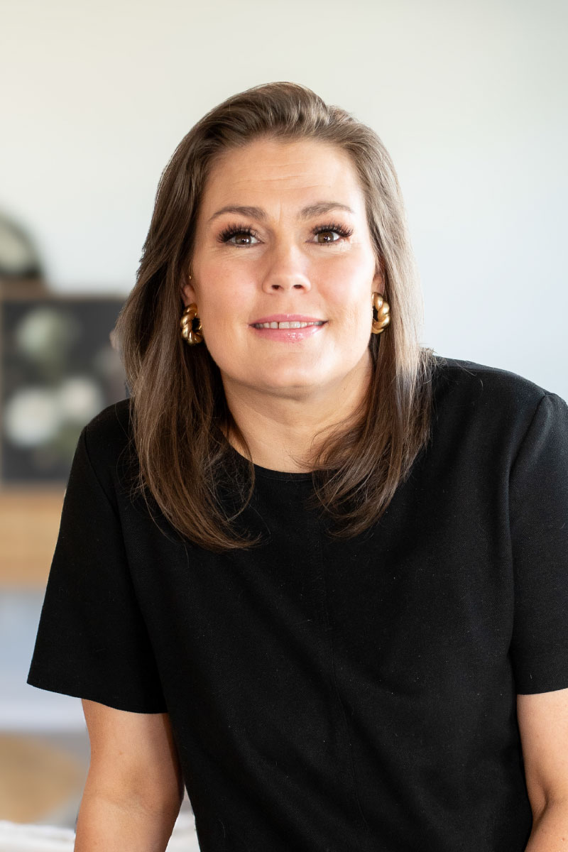 Professional headshot of a confident woman with straight shoulder-length brown hair, wearing a black top and gold hoop earrings, posing indoors with a soft, modern background.