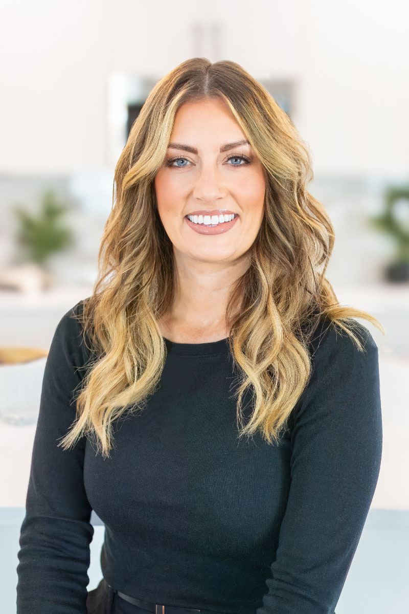 Professional headshot of Jody Goedhart with long, styled blonde hair wearing a black top, photographed in a bright, modern interior setting with a soft, blurred background.