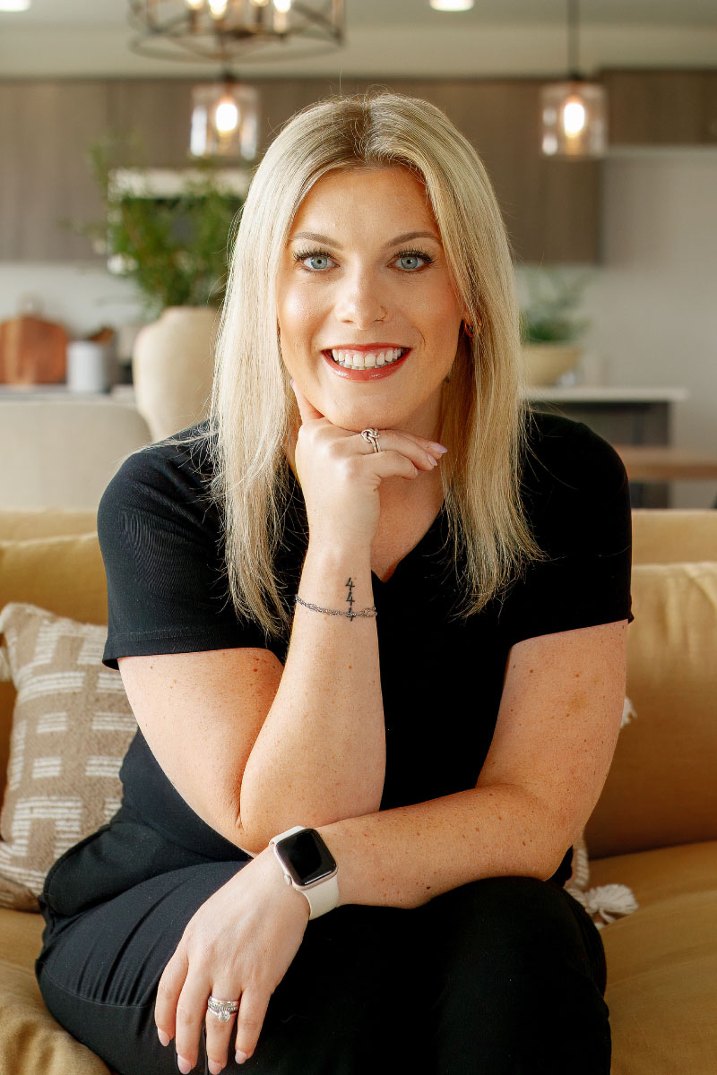 Smiling woman with blonde hair wearing a black outfit, seated on a tan couch in a modern home interior.
