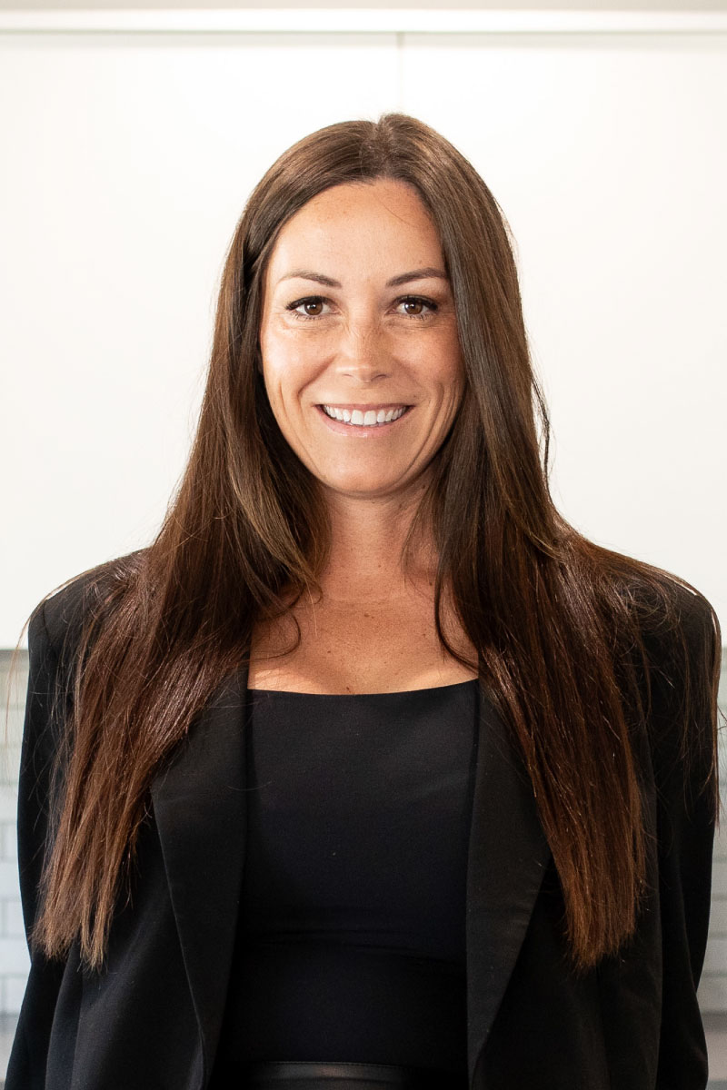 Professional headshot of a smiling woman with long straight brown hair, dressed in a black blazer and top, standing in a bright modern interior with a white background.