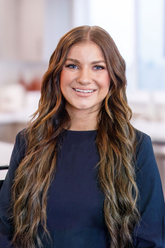 Professional headshot of Macy Campbell, an agent at CBH Homes, smiling with long brunette hair in a bright, softly blurred interior setting