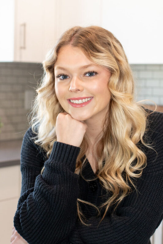 Professional headshot of a smiling young woman with long blonde wavy hair, wearing a black knit sweater, resting her chin on her hand, posed in a bright modern kitchen with white cabinetry and gray tile backsplash.