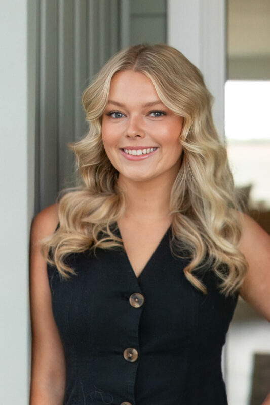 Smiling woman with blonde wavy hair wearing a black sleeveless top, professional headshot.
