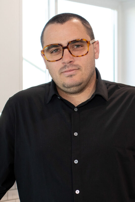 Professional headshot of a man with short dark hair wearing tortoiseshell glasses and a black button-up shirt, standing in a well-lit modern interior with natural light from large windows.