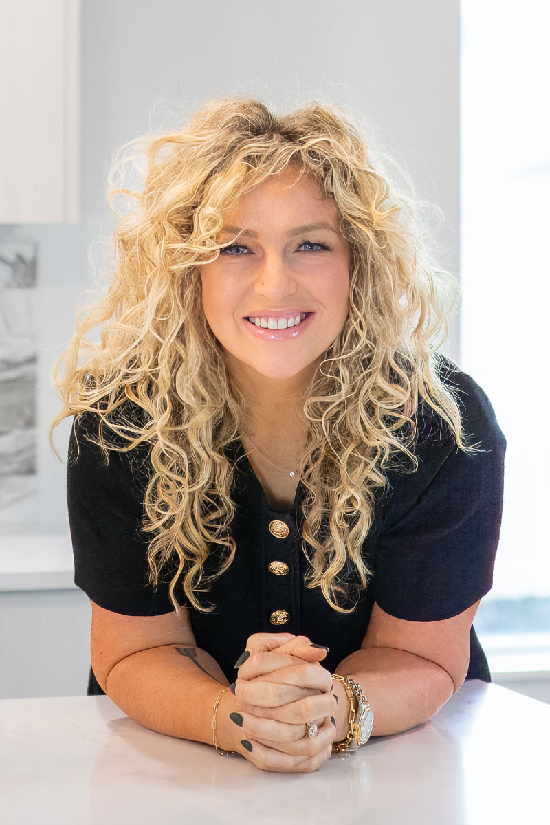Professional headshot of Teschia Tucakovic smiling with curly blonde hair in a modern, bright setting