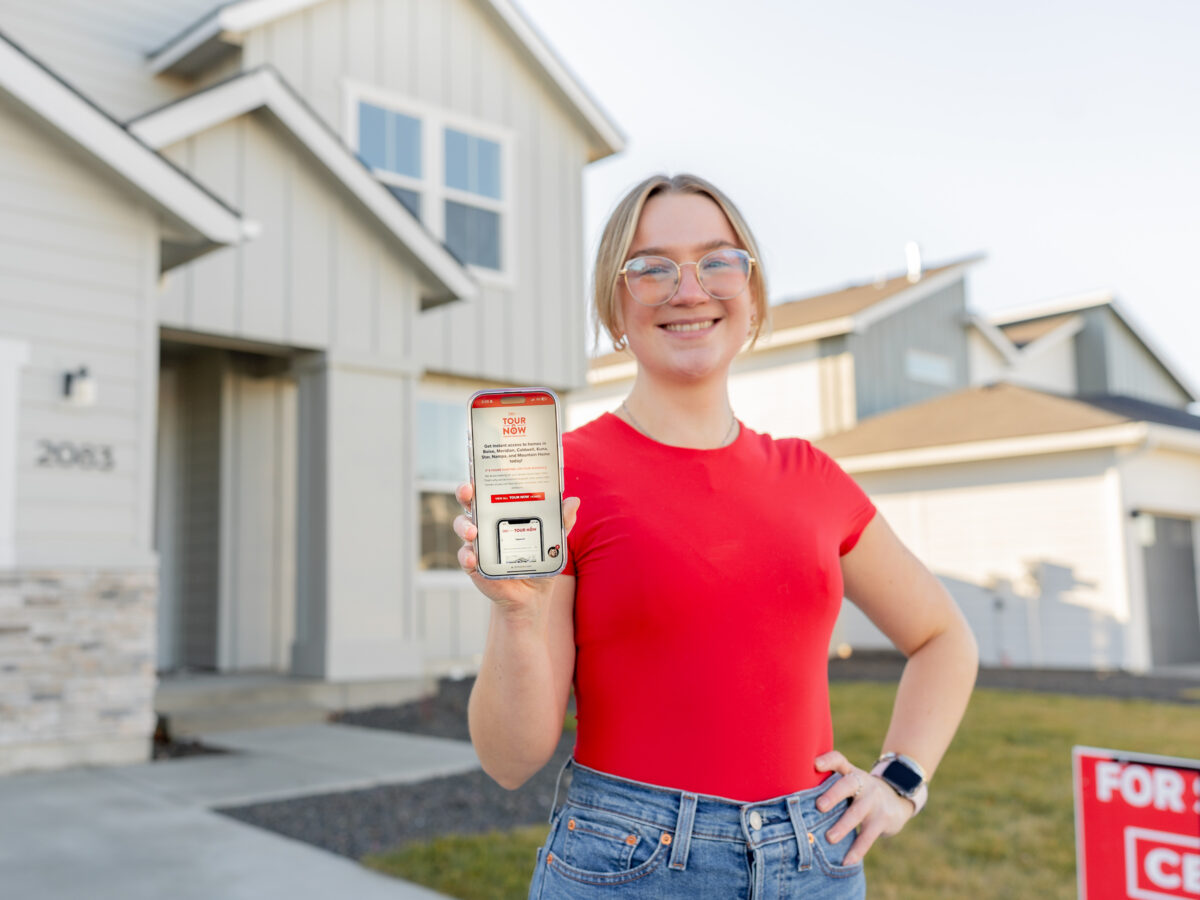 A smiling person stands in front of a modern CBH Homes house, holding a smartphone displaying the “Tour Now” page, promoting CBH Homes’ self-guided home tours.