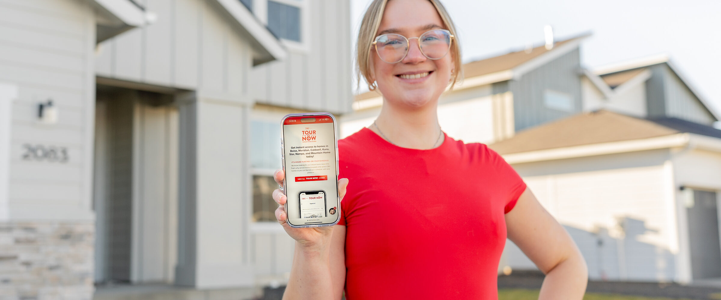 A smiling person stands in front of a modern CBH Homes house, holding a smartphone displaying the “Tour Now” page, promoting CBH Homes’ self-guided home tours.