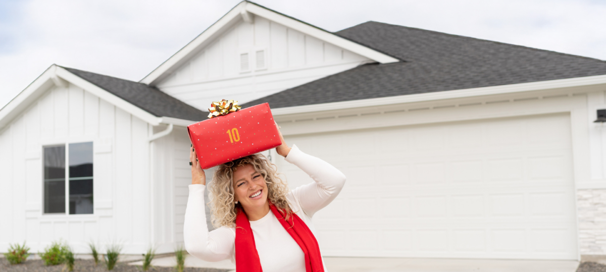 Smiling woman holding a red gift box labeled “10” in front of a white CBH model home during the 10th Annual Festival of Homes.