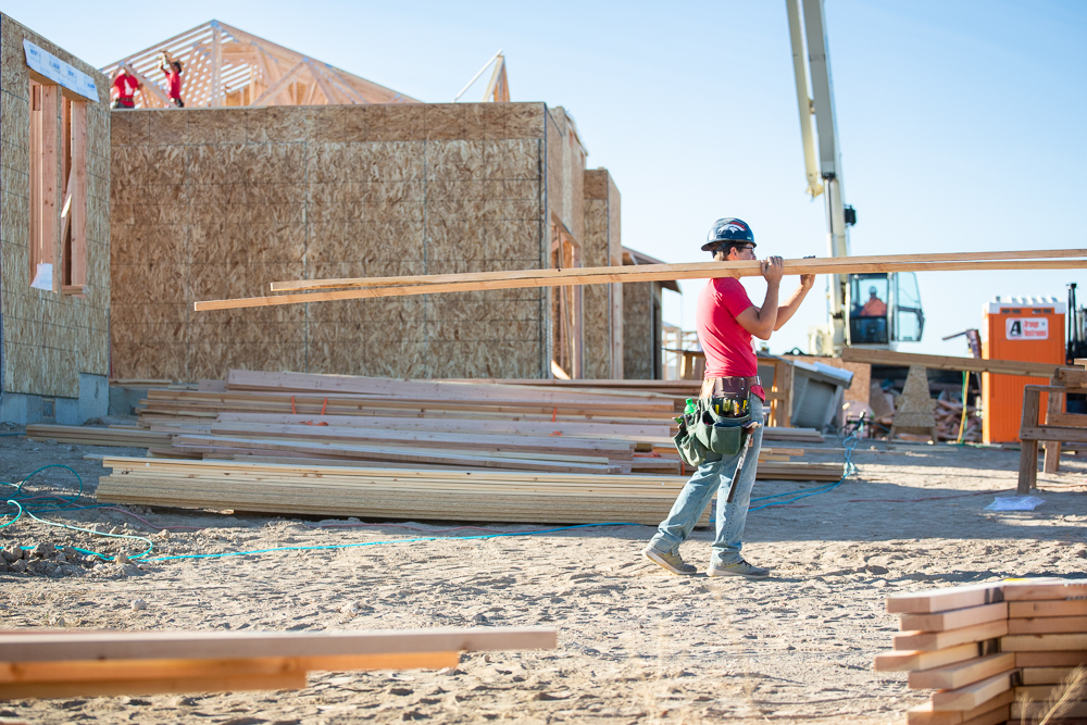 Construction worker carrying wooden beams at a new home building site in Idaho with framing and scaffolding in progress.