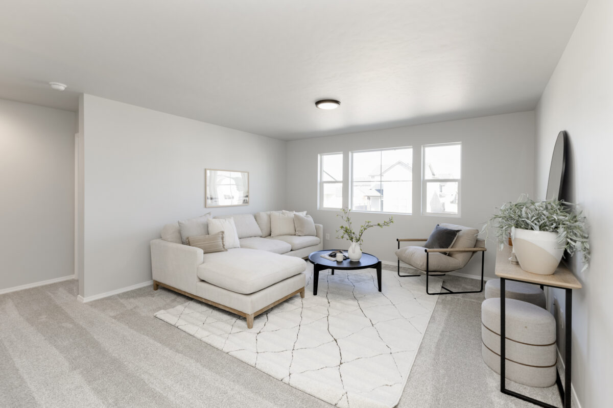 Bright and airy living room with neutral-toned decor, featuring a large L-shaped sectional sofa, a modern lounge chair, and a black round coffee table on a patterned area rug. Three large windows allow natural light to fill the space, while a minimalist console table with greenery and ottomans adds style and functionality. The soft gray carpet and light walls create a serene and cozy atmosphere.