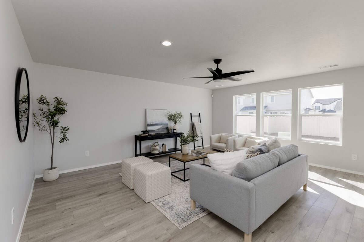 "Spacious and sunlit living room featuring a modern gray sofa, two white accent chairs, and a wooden coffee table on a patterned area rug. Three large windows bring in natural light, illuminating the light wood flooring. The room is accented with minimalist decor including a black ceiling fan, potted plants, and a sleek black console table with a landscape painting and books. Neutral tones and clean lines create a calm, inviting atmosphere.