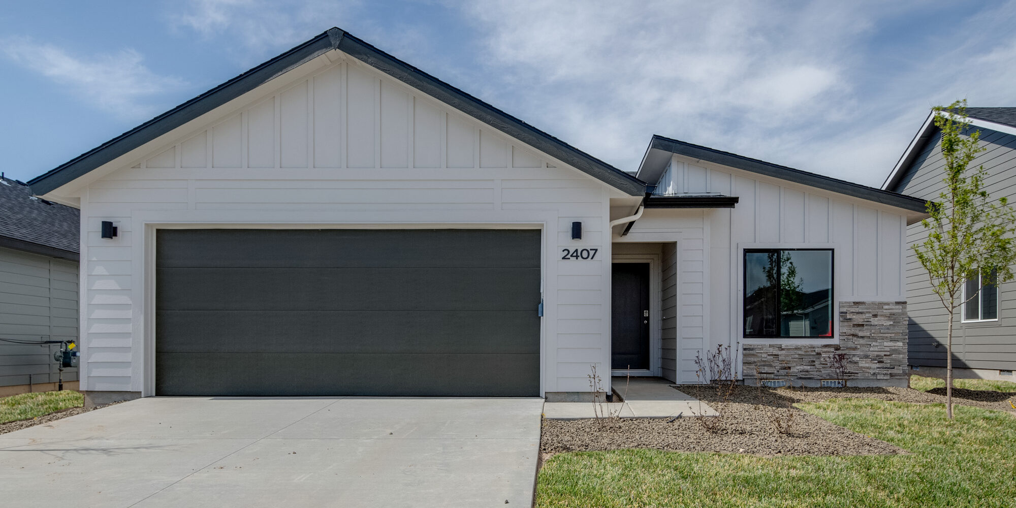 Front exterior view of a modern single-story house with white vertical siding and dark trim, featuring a two-car garage with a dark door, a stone accent wall around a large front window, a small front porch, and a landscaped front yard with young trees and fresh sod.