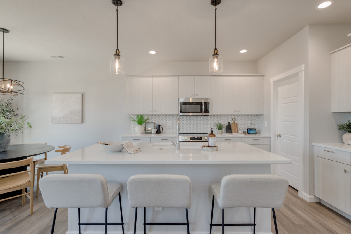 Bright and modern kitchen with a large white quartz island featuring an undermount sink and three upholstered barstools. White cabinetry with sleek hardware lines the back wall, paired with a subtle geometric backsplash and stainless steel appliances. Pendant lights with clear glass shades hang above the island, and a cozy dining area with wooden chairs and a black round table is visible to the left.