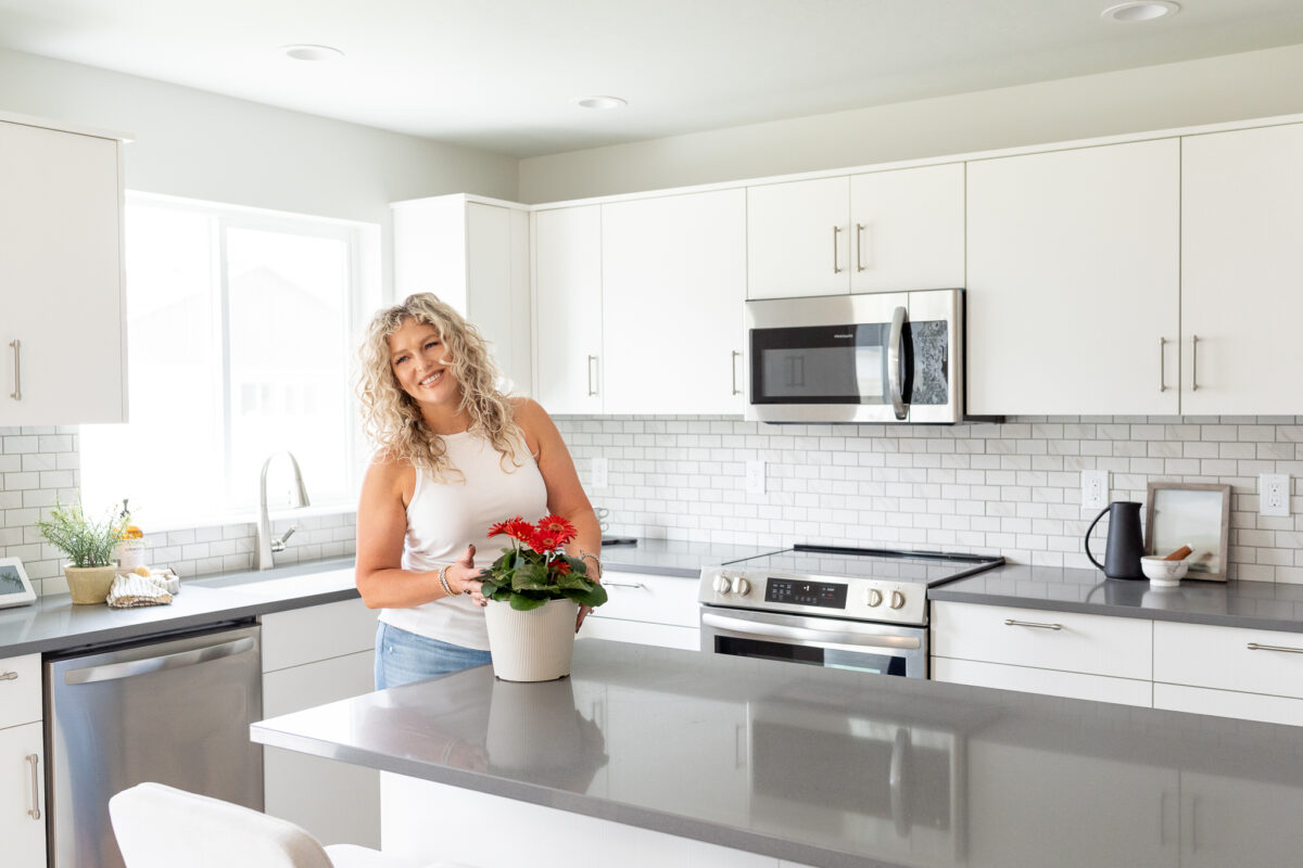 Smiling woman with curly blonde hair standing in a bright modern kitchen with white cabinets, gray countertops, and stainless steel appliances, holding a white flower pot with red flowers on a central island.