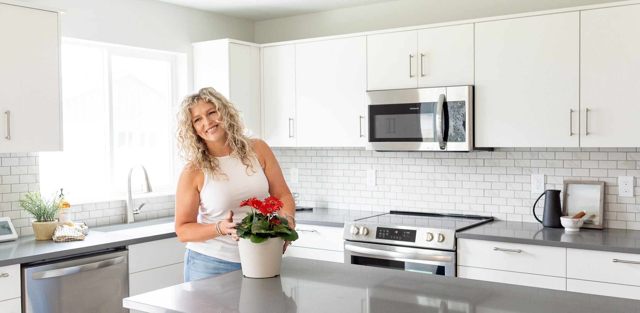 Smiling woman with curly blonde hair standing in a bright modern kitchen with white cabinets, gray countertops, and stainless steel appliances, holding a white flower pot with red flowers on a central island.