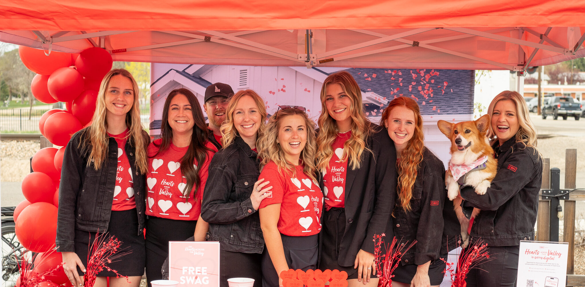 A group of eight smiling CBH Homes team members, all wearing red “Hearts for the Valley” shirts and black jackets, pose together under a red tent decorated with red balloons. They stand behind a red table offering free swag, informational flyers, and decorations with heart motifs. One team member holds a corgi dog wearing a bandana. The atmosphere is cheerful and welcoming, likely at a community event or outreach gathering.
