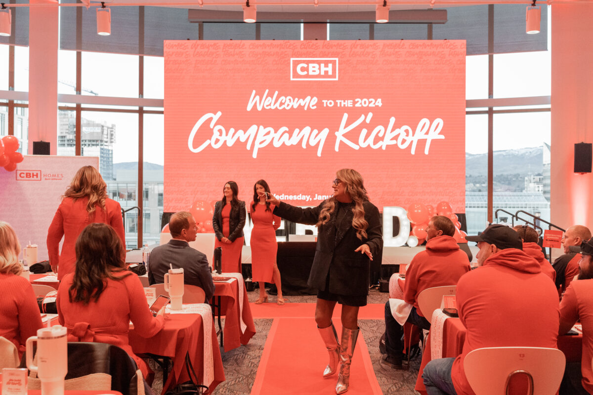 CBH Homes 2024 Company Kickoff event with employees dressed in red seated at decorated tables, a large welcome screen in the background, and a woman in silver boots speaking down the center aisle.