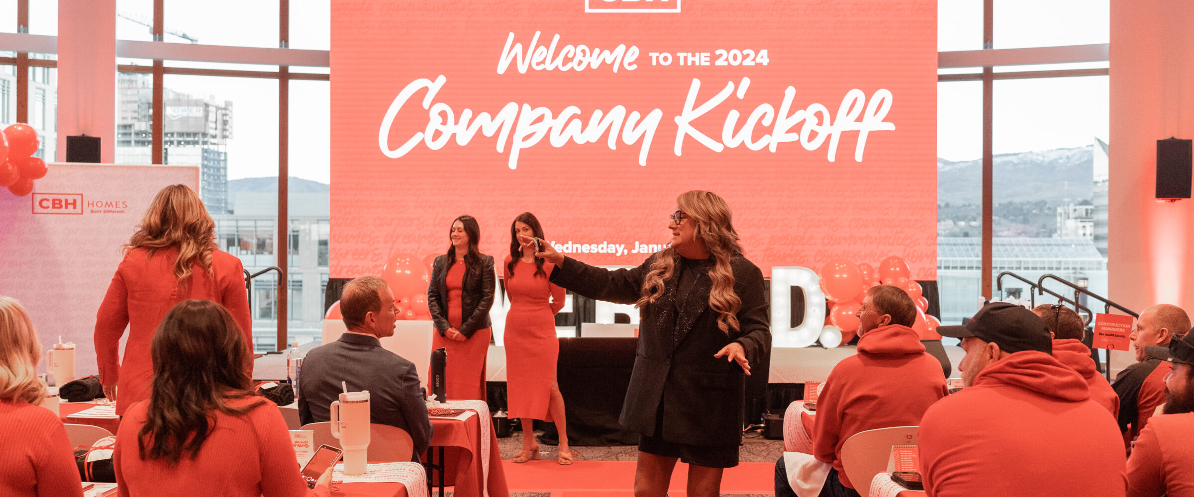 CBH Homes 2024 Company Kickoff event with employees dressed in red seated at decorated tables, a large welcome screen in the background, and a woman in silver boots speaking down the center aisle.