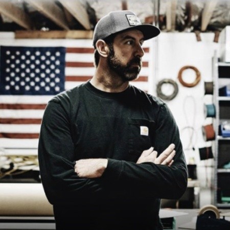 Artist Chris Foster of FosterWeld standing in his workshop with folded arms, wearing a cap and dark shirt, with an American flag hanging in the background.