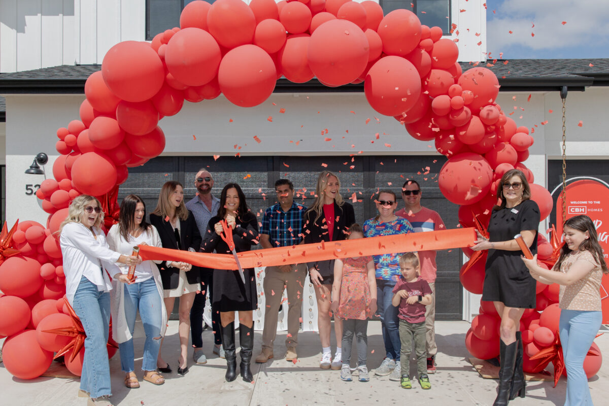 CBH Homes team and community members celebrate a grand opening with a ribbon-cutting ceremony under a red balloon arch.