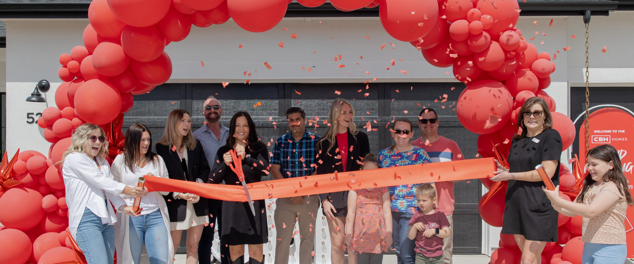 CBH Homes team and community members celebrate a grand opening with a ribbon-cutting ceremony under a red balloon arch.