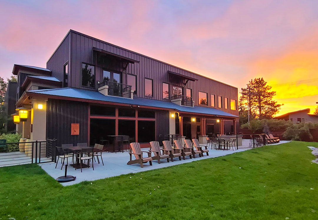 Exterior view of Telaya Wine Co. at sunset with modern architecture, patio seating, and lush green lawn overlooking the Boise River.
