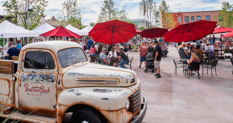 Crowds gather under red umbrellas at Indian Creek Plaza in Caldwell, Idaho, during a community event, with a vintage white truck featuring the Indian Creek Plaza logo in the foreground.