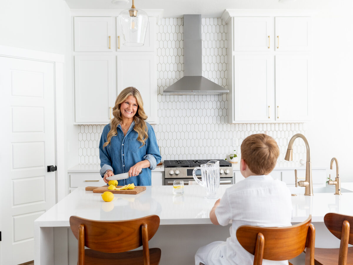 A woman with long blonde wavy hair, wearing a denim shirt, smiles while slicing lemons on a wooden cutting board in a bright, modern kitchen. A young boy with short red hair, dressed in a white shirt, sits at the kitchen island on a wooden chair, watching her. A glass pitcher of lemonade and a drinking glass are on the white countertop. The kitchen features white cabinets, gold hardware, a stainless steel range hood, and a geometric white tile backsplash.