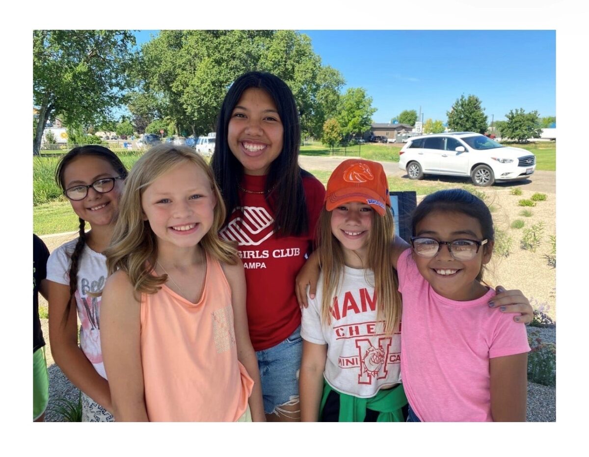 A group of five smiling girls, including a Boys & Girls Club of Canyon County team member in a red club shirt, enjoying a sunny day outdoors. They stand close together, radiating joy and connection, with trees, landscaping, and a white SUV in the background.