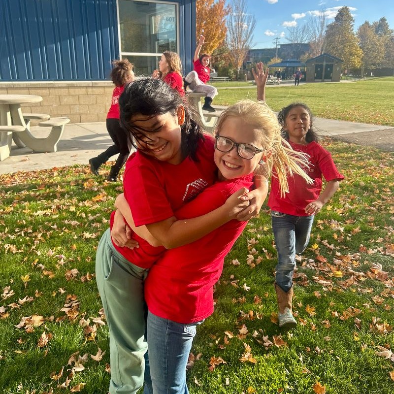 Two young girls wearing red shirts hug and smile while playing outside on a sunny autumn day, surrounded by fallen leaves and other children laughing and running in the background.