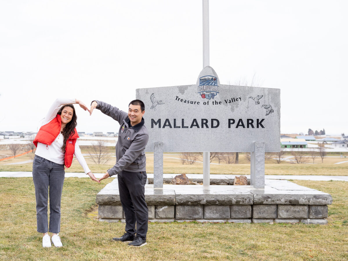 Two smiling people standing in front of the Mallard Park sign in Caldwell, Idaho, forming a heart shape with their arms. The background shows an open grassy area with houses in the distance.
