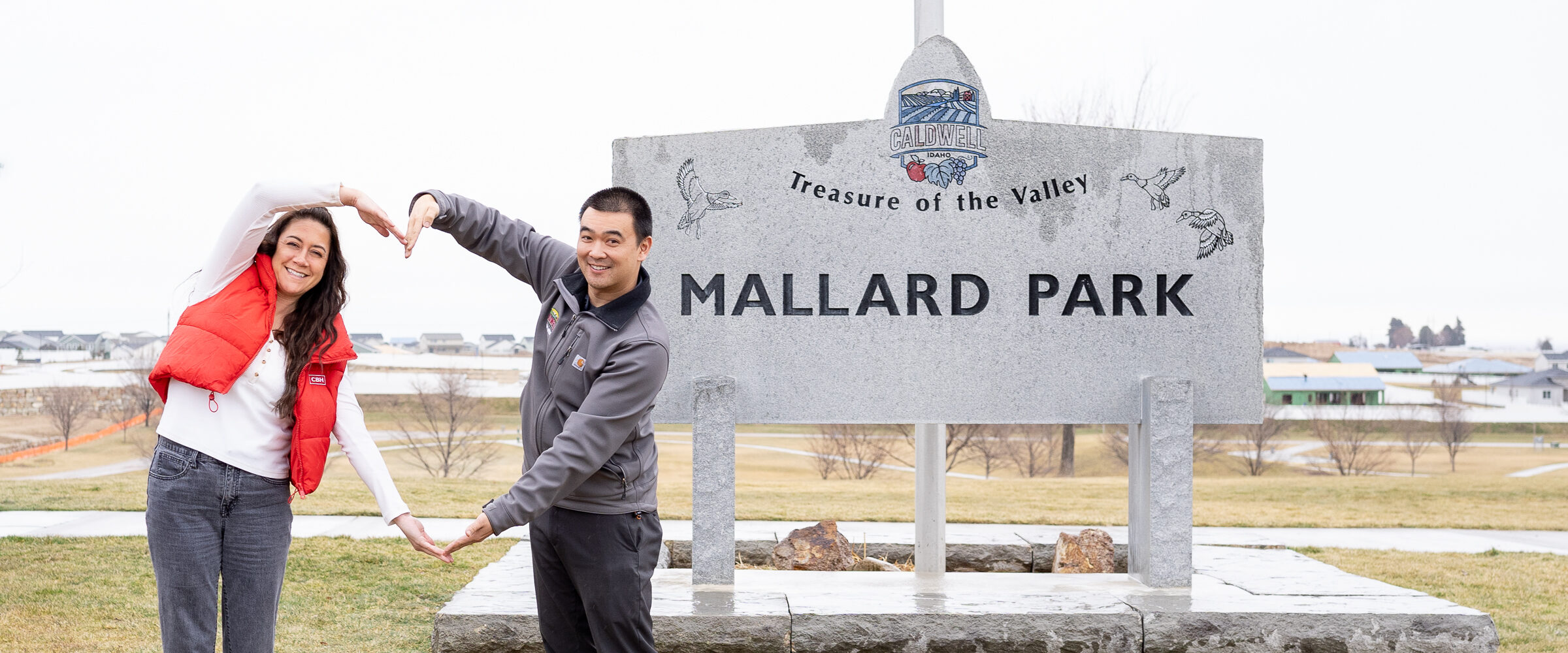 Two smiling people standing in front of the Mallard Park sign in Caldwell, Idaho, forming a heart shape with their arms. The background shows an open grassy area with houses in the distance.