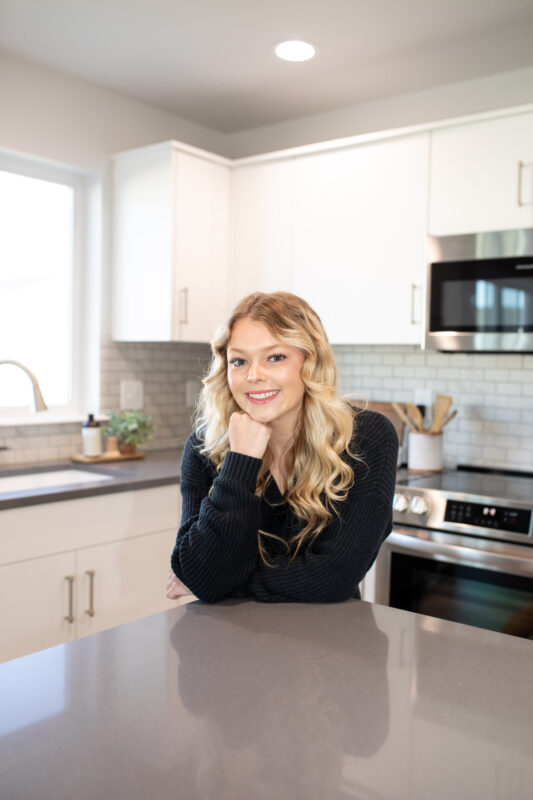Smiling woman with long blonde hair in dark sweater standing in modern kitchen featuring white cabinets, stainless steel appliances, and quartz countertops.