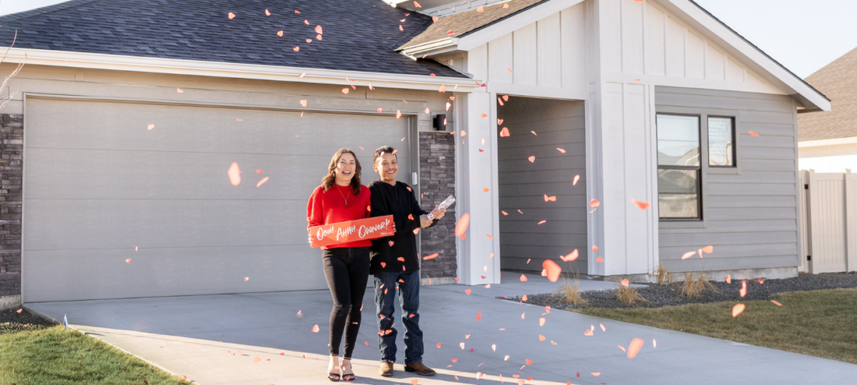 Happy new homeowners celebrating in front of their modern gray house with confetti and a sign that says ‘Oooh! Ahhh! Owner!'