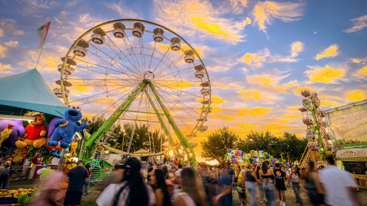 A lively fairground at sunset with a large Ferris wheel as the main attraction. The sky is filled with vibrant orange and yellow clouds. People are walking around, enjoying games and rides, and there are colorful tents and booths, including one with large stuffed toys on display.