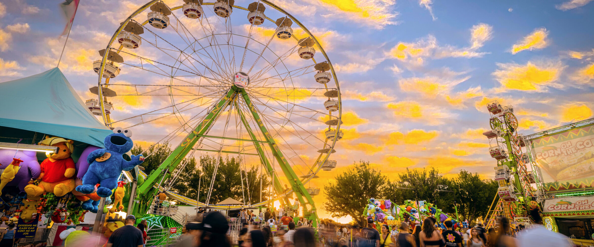 A lively fairground at sunset with a large Ferris wheel as the main attraction. The sky is filled with vibrant orange and yellow clouds. People are walking around, enjoying games and rides, and there are colorful tents and booths, including one with large stuffed toys on display.