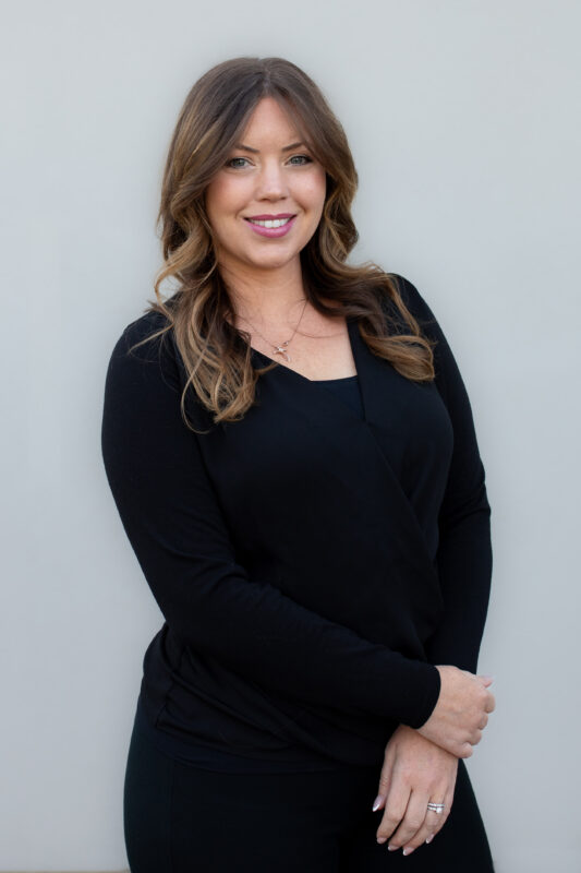 Professional headshot of a smiling woman with long, styled brown hair wearing a black blouse, standing against a white background.