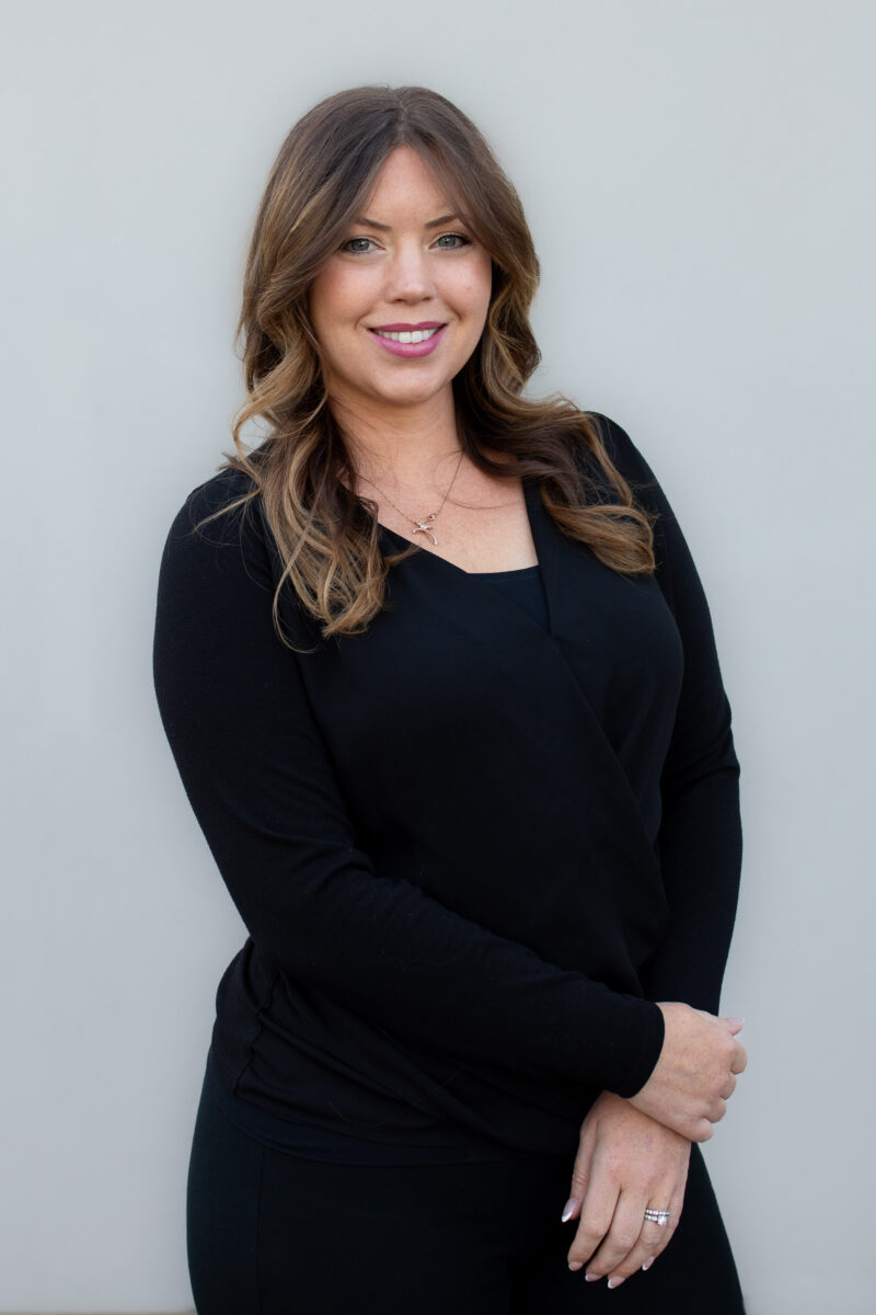 Professional headshot of a smiling woman with long, styled brown hair wearing a black blouse, standing against a white background.