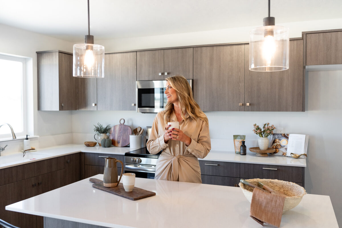 Woman standing in a bright, modern kitchen with dark wood cabinets and white countertops, holding a coffee cup and smiling beside the island with decor accents.